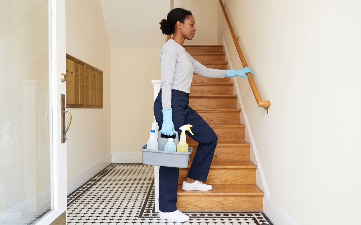 A cleaner from an apartment cleaning service steps onto the wood stairs of a building's front hall with her caddy, one hand on the rail, black and white mosaic tile underfoot.