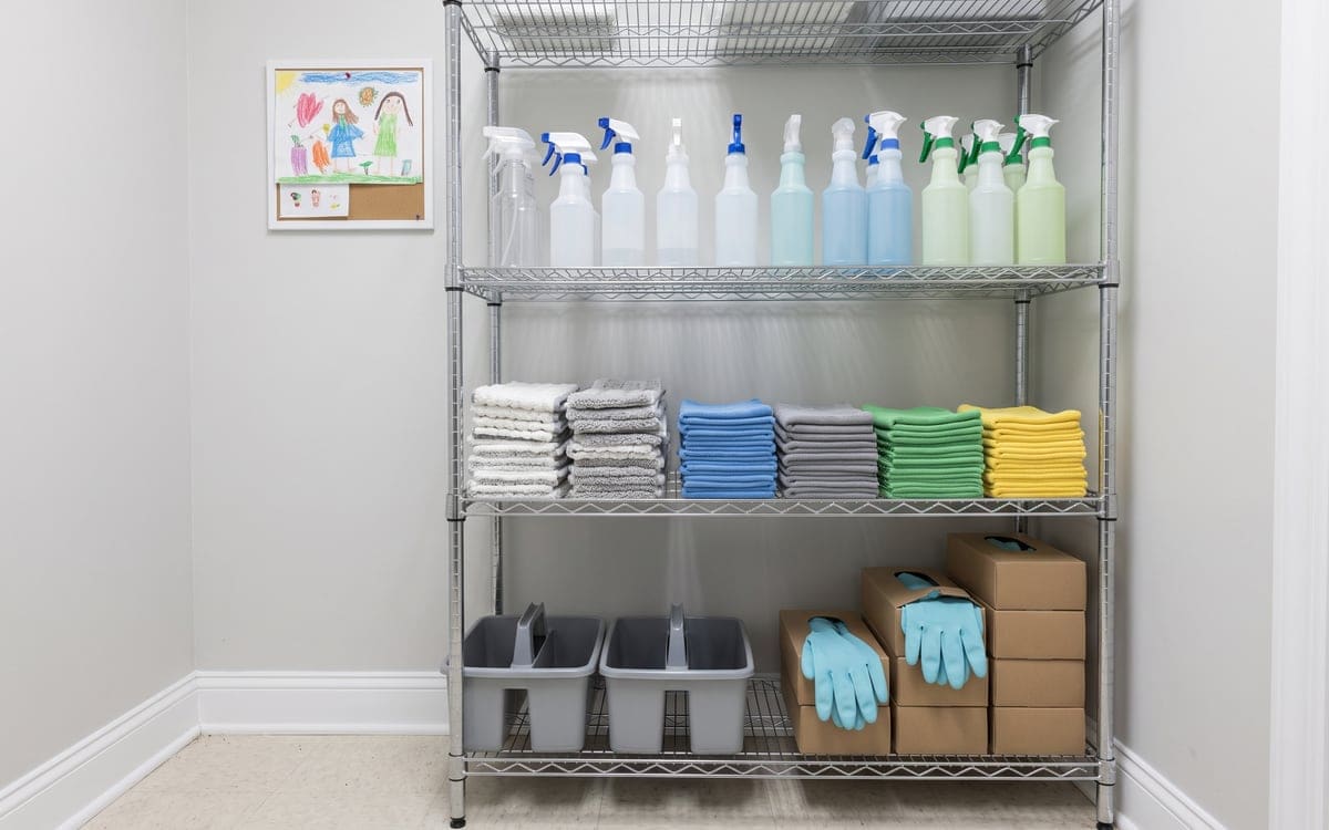 A house cleaning company's wire supply shelf, microfiber cloths stacked by color under a row of spray bottles, with two gray caddies and boxes of blue gloves on the bottom.