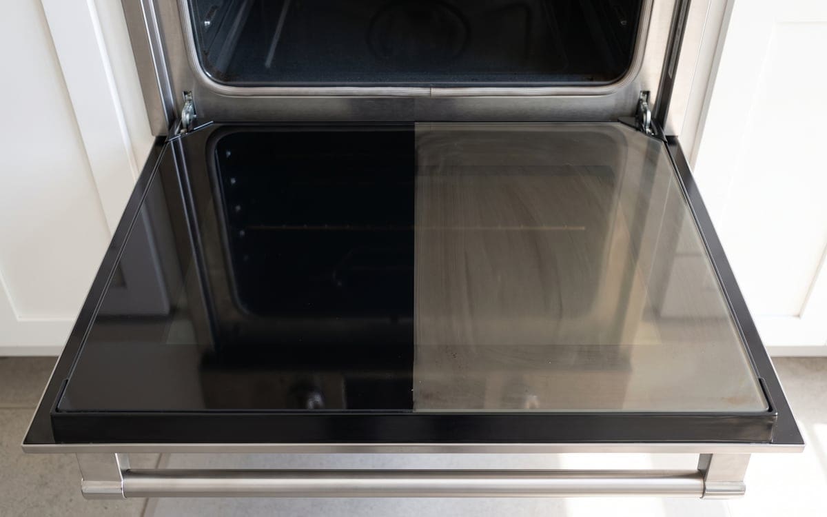 An oven door lowered open for deep cleaning service. The left half of its glass is wiped glossy black and the right half still has a brown film.