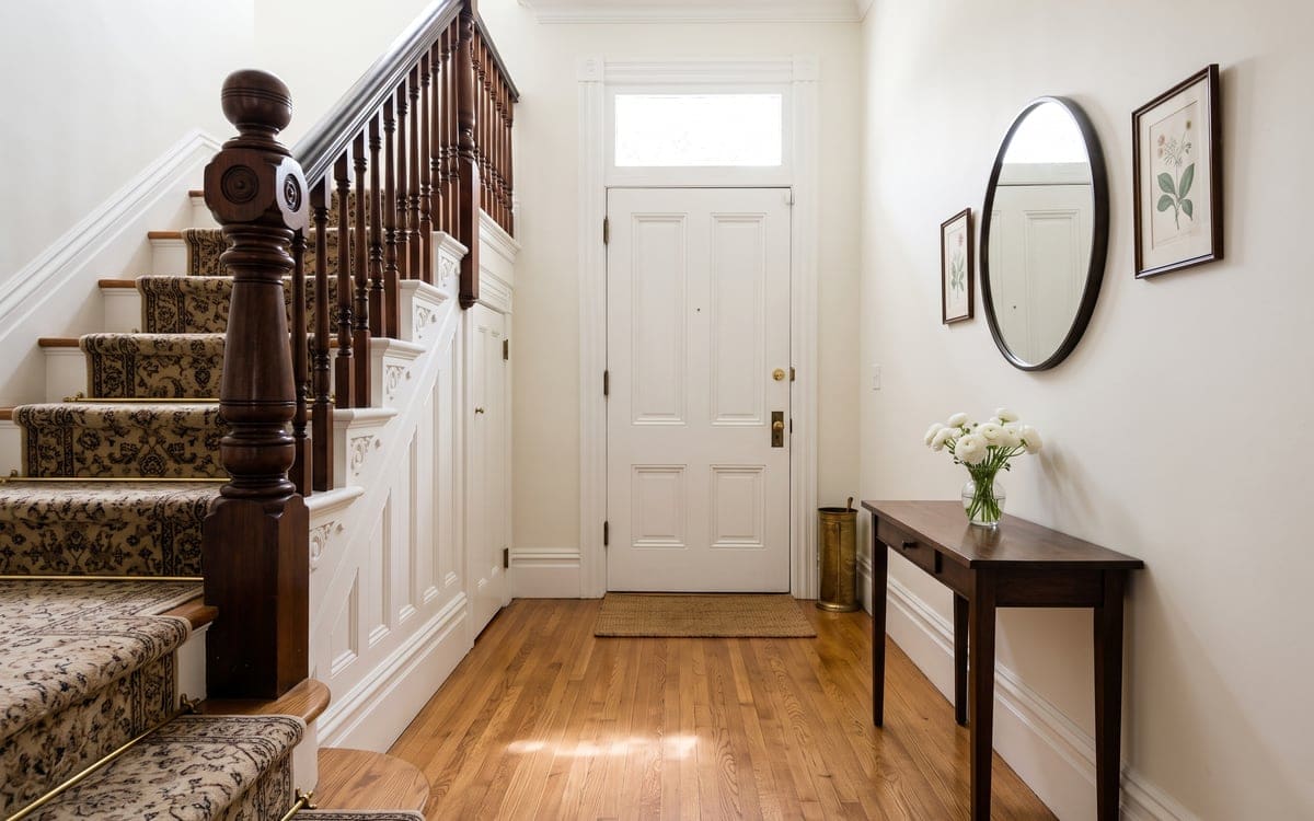 A front hall after cleaning services, its oak floor shining between a staircase with a patterned runner and brass stair rods and a dark console table holding white flowers.