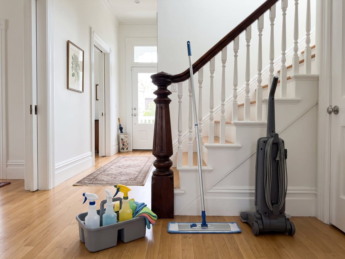 A San Francisco front hall set up for a house cleaning. A caddy of spray bottles waits on the oak floor, a mop leans by the newel post and an upright vacuum stands at the foot of the stairs.