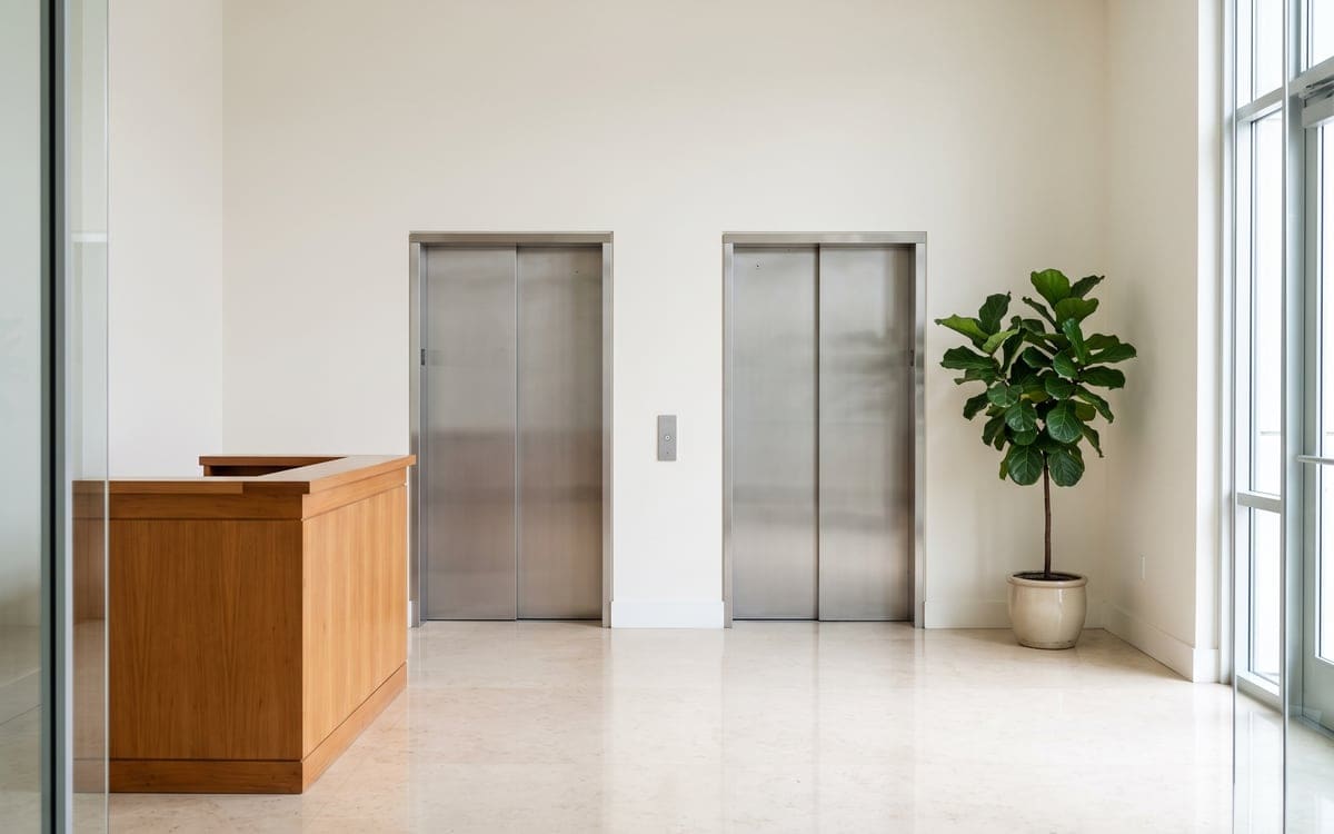 A condo lobby with an empty wood front desk and two steel elevators, a potted fiddle-leaf fig standing in the corner.