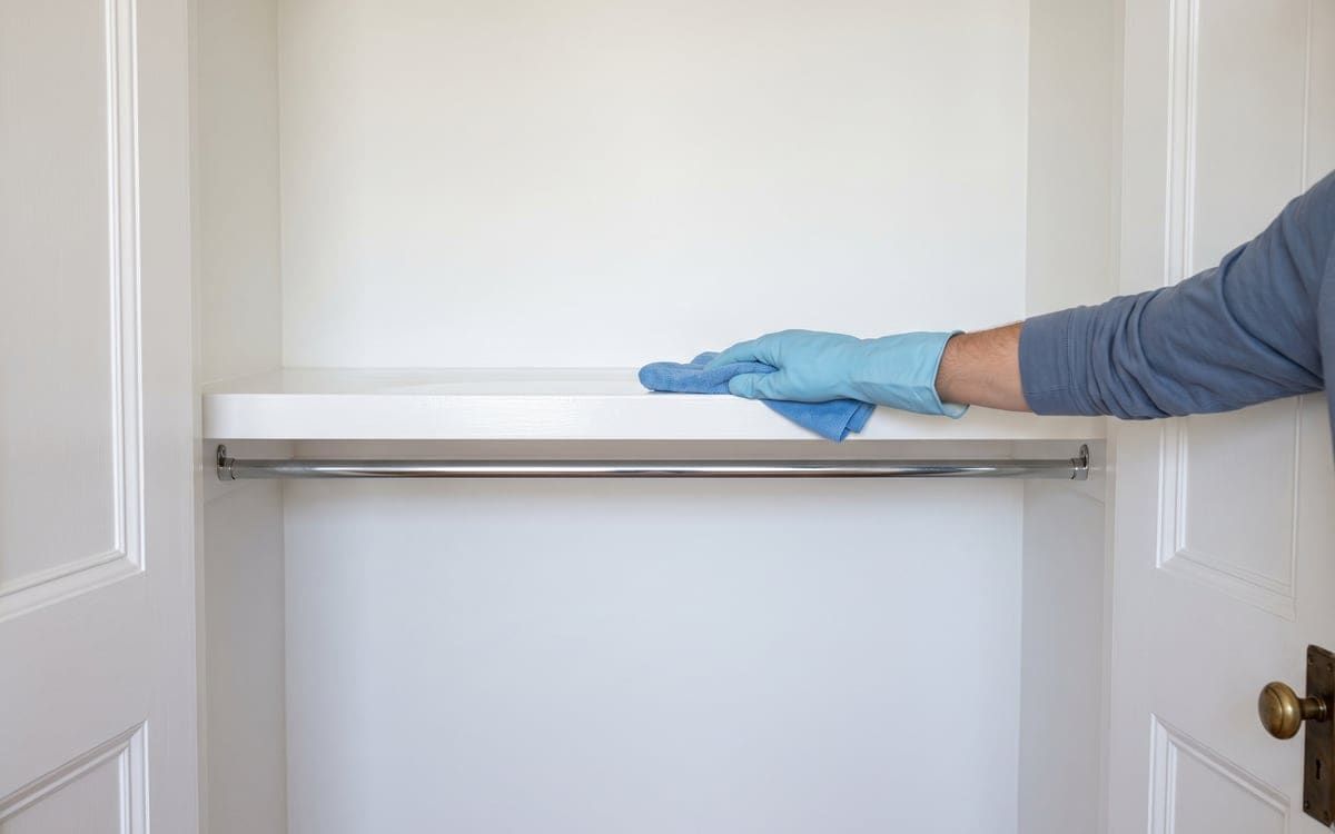A gloved hand wipes the white shelf of an empty closet with a blue cloth during move out cleaning, above a bare hanging rod.
