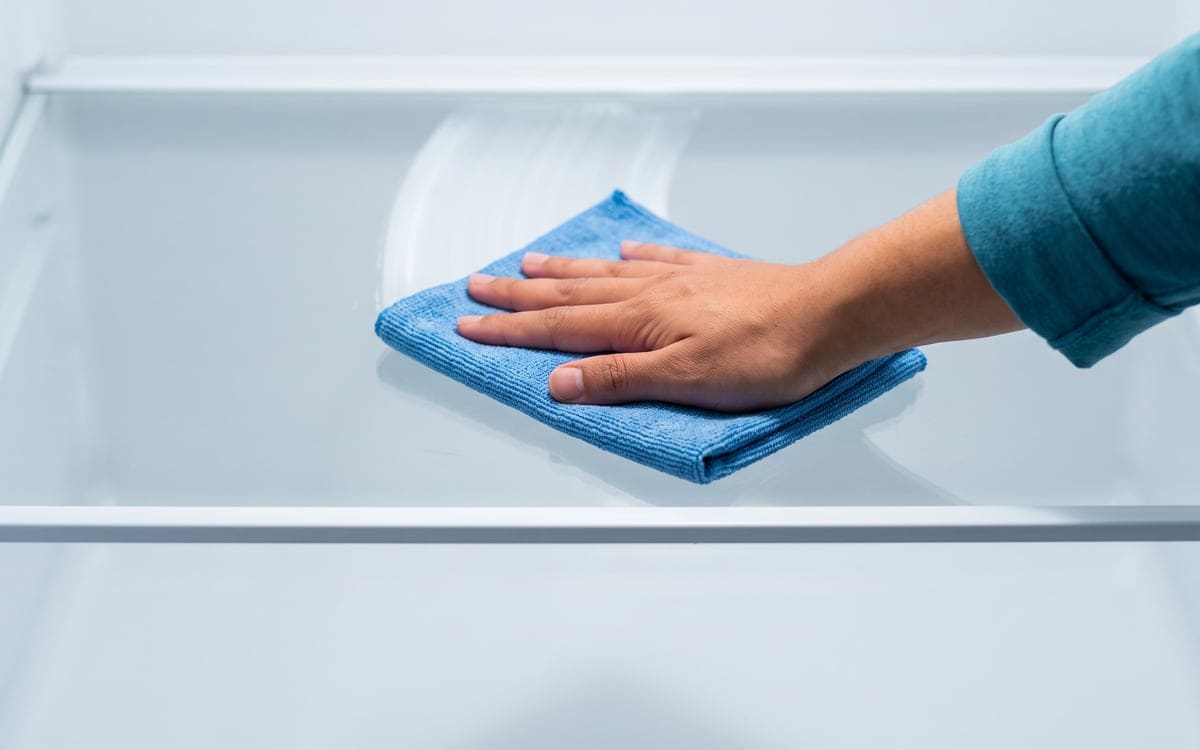 A bare hand in a teal sleeve wipes an empty glass refrigerator shelf with a folded blue cloth.