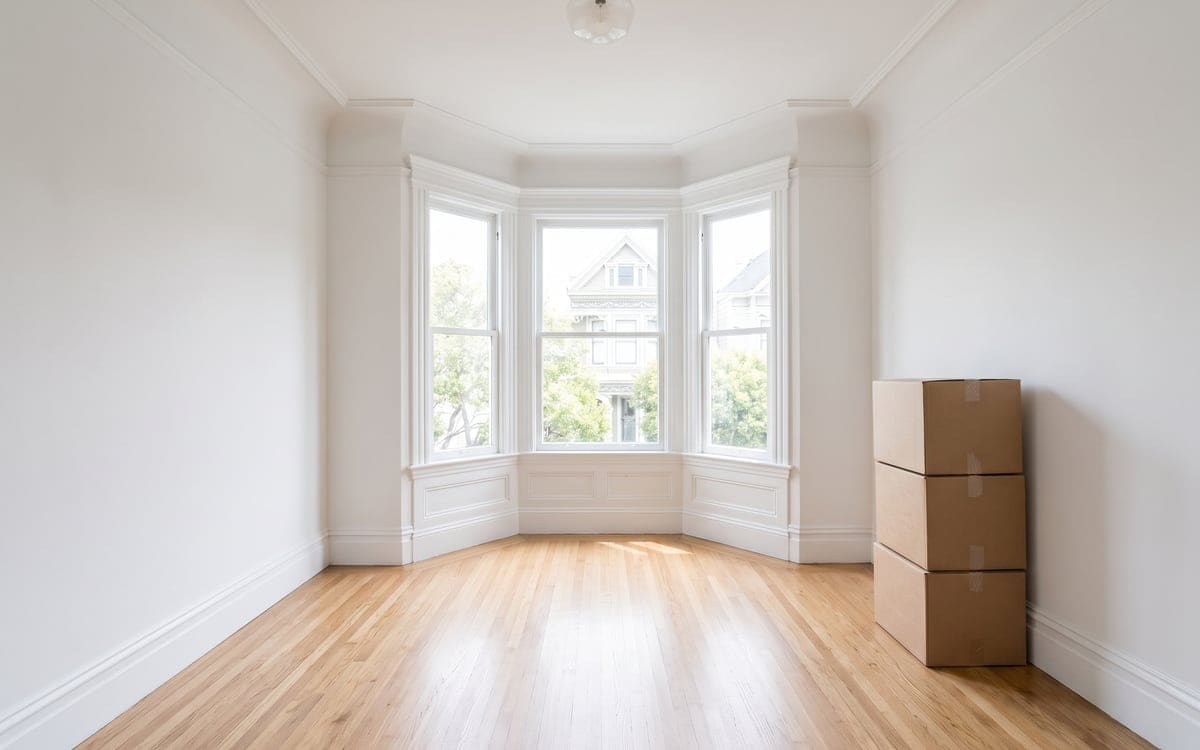 An empty San Francisco bedroom on move-out day, three taped boxes stacked against the wall and the oak floor bare under a bay window.