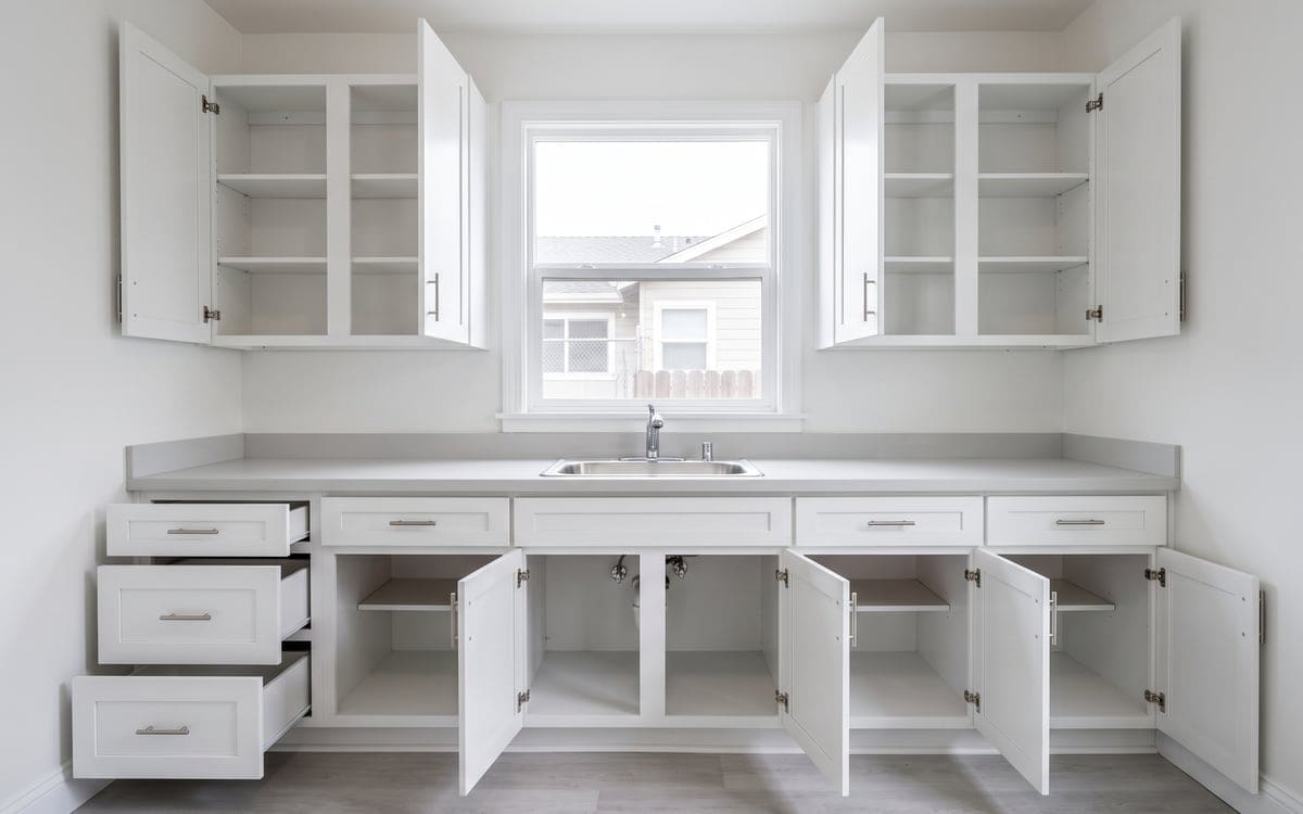 Every cabinet door and drawer stands open on bare white shelves along the sink wall of an empty kitchen after move out cleaning.