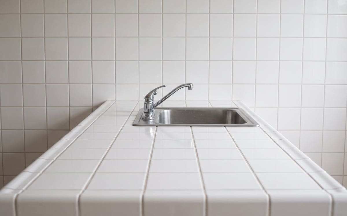 A bare white tile counter and a dry stainless sink with a chrome lever faucet, set against a white tile backsplash.
